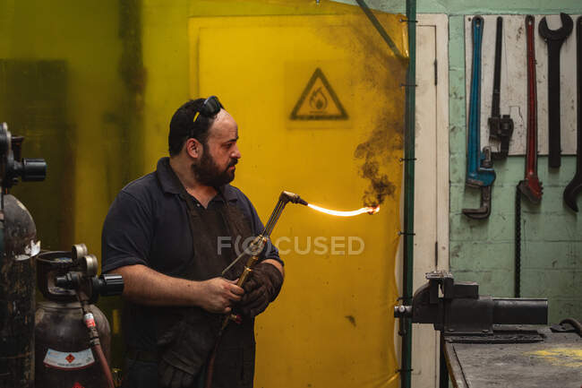 Trabajador de fábrica masculino caucásico que usa delantal oscuro y guantes de seguridad, de pie en un banco de trabajo y soldadura. - foto de stock