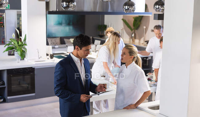Caucasian female chef wearing chefs whites talking with a smartly dressed mixed race male colleague holding a clipboard in a modern restaurant kitchen, with other kitchen staff busy in the background — Stock Photo