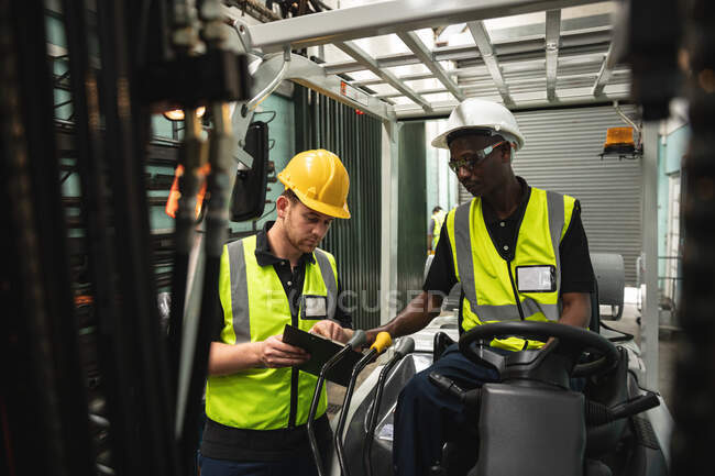 Caucasian and African American male factory workers wearing a high vis vest talking, holding clipboard and operating machinery. Workers in industry at a factory making hydraulic equipment. — Stock Photo