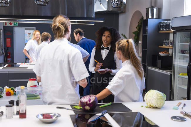 Mixed Race Kellnerin, schreibt auf ein Notizbuch, spricht mit zwei kaukasischen Köchinnen, während andere Köche im Hintergrund stehen. Kochkurs in einer Restaurantküche. — Stockfoto