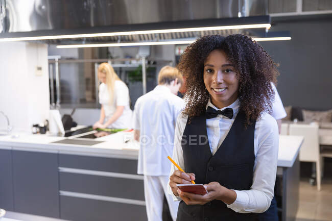 Portrait d'une serveuse métissée écrivant sur un carnet, regardant la caméra et souriant, avec des chefs cuisiniers cuisiner et couper des légumes en arrière-plan. Cours de cuisine dans une cuisine de restaurant. — Photo de stock