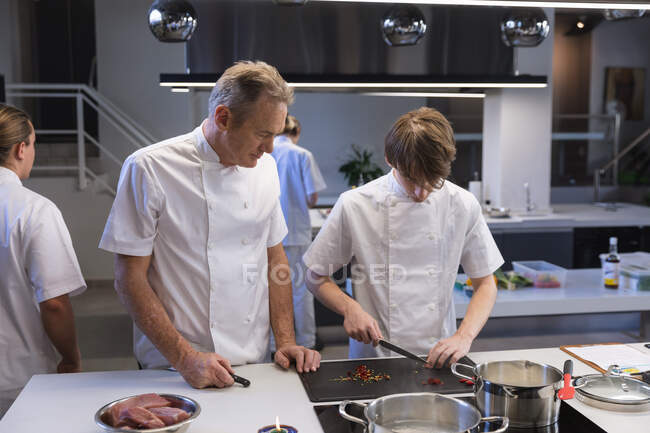 Junger kaukasischer Koch schneidet Zutaten für das Kochen, während der leitende kaukasische Koch neben ihm steht und auf seine Hände schaut, während andere Köche im Hintergrund kochen. — Stockfoto