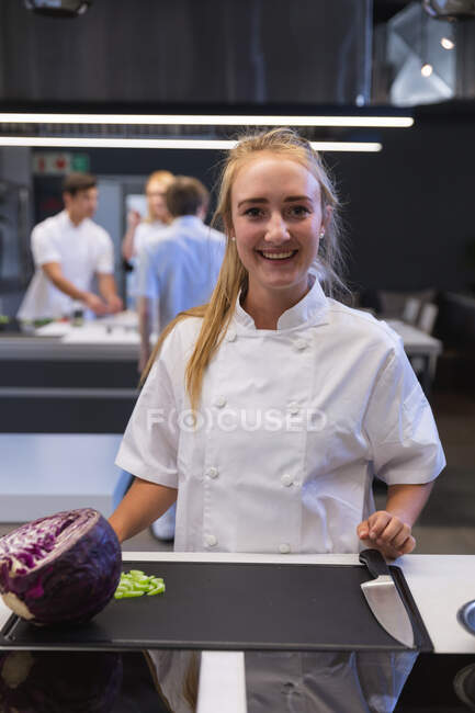 Portrait of a Caucasian female chef looking at the camera and smiling, with other chefs cooking in the background. Cookery class at a restaurant kitchen. — Stock Photo