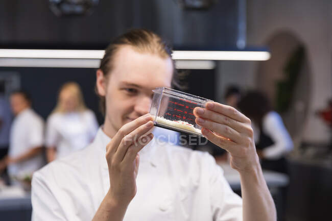 Caucasian male chef looking at cooking ingredients in a plastic box and smiling, with other chefs cooking in the background. Cookery class at a restaurant kitchen. — Stock Photo