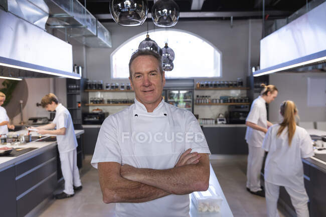 Portrait of a senior Caucasian male chef, crossing his arms, looking at the camera and smiling with other chefs cooking in the background. Cookery class at a restaurant kitchen. — Stock Photo