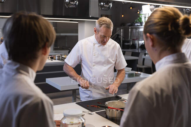 Senior Caucasian chef cutting vegetables, with other chefs watching in the foreground. Cookery class at a restaurant kitchen. — Stock Photo