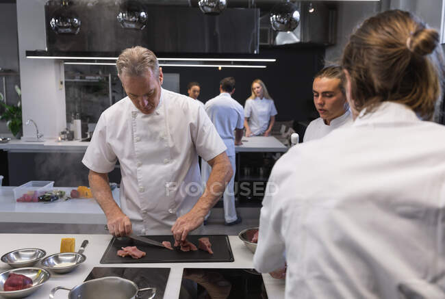 Chef caucasien principal coupant des légumes, avec d'autres chefs regardant au premier plan. Cours de cuisine dans une cuisine de restaurant. — Photo de stock