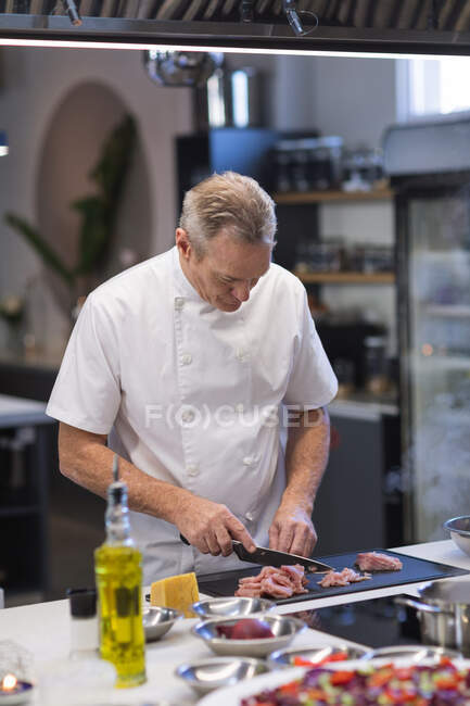 Старший кавказький шеф-кухар стоїть біля столу, зрізає шматок риби, дивлячись на нього. Клас куховарства на кухні.. — стокове фото