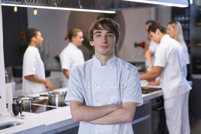 Portrait of a Caucasian male chef crossing his arms, looking at the camera and smiling, with other chefs cooking in the background. — Stock Photo