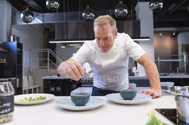 Lo chef maschio caucasico più anziano versa il sale su un piatto pronto, sorridendo. Classe di cucina in una cucina ristorante. — Foto stock