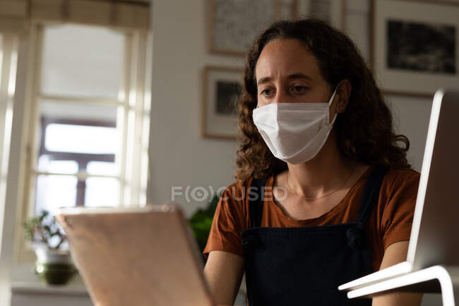 Mujer caucásica pasando tiempo en casa, trabajando desde casa, usando una máscara facial. Estilo de vida en el hogar aislado en cuarentena durante la pandemia del coronavirus covid 19. - foto de stock