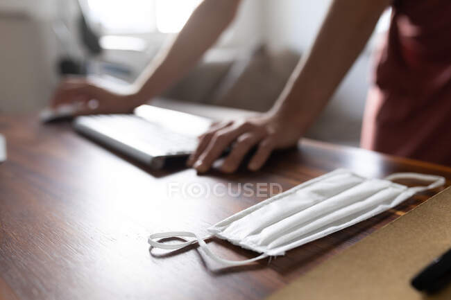 Hands of woman spending time at home, using her computer, face mask on the desk. Lifestyle at home isolating, social distancing in quarantine lockdown during coronavirus covid 19 pandemic. — Stock Photo