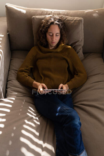 Белая женщина проводит время дома, пользуется планшетом, отдыхает на диване. Стиль жизни дома изолирует, социальное дистанцирование в карантинной изоляции во время пандемии коронавируса ковид 19. — стоковое фото