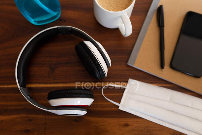 Close up of a face mask, hand sanitizer, a mug with coffee and headphones on a wooden desk. Lifestyle at home isolating, social distancing in quarantine lockdown during coronavirus covid 19 pandemic. — Stock Photo