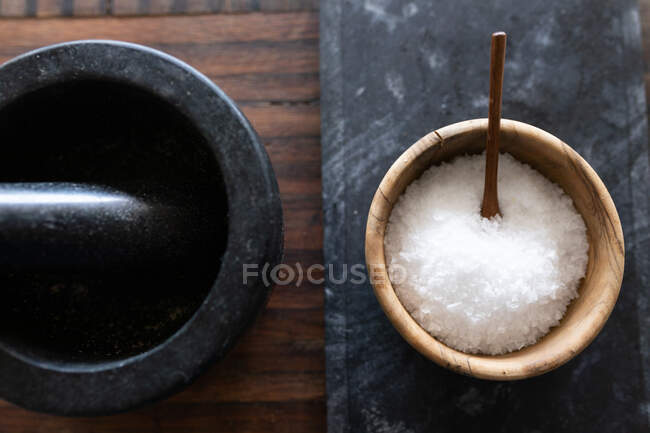 Close up of a mortar and salt in a container on wooden and graphite table. Food preparation at home promoting healthy lifestyle. — Stock Photo