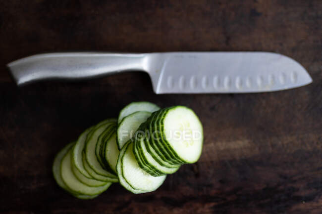 Overhead close up of a sharp knife and slices of cucumber on a wooden table. Food preparation at home promoting healthy lifestyle. — Stock Photo