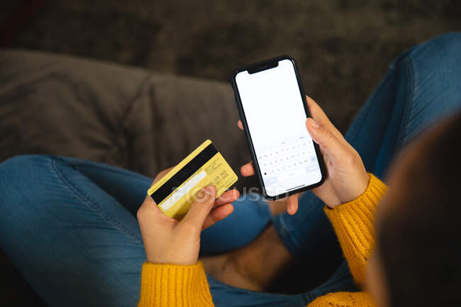 Vista aérea de la mujer sentada en un sofá en casa, con una tarjeta de crédito y un teléfono inteligente, pagando en línea. Comercio electrónico conveninece compras. - foto de stock