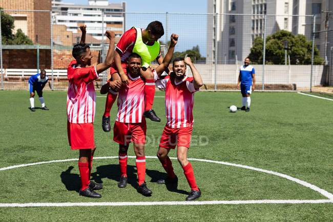Zwei multiethnische Mannschaften von fünf männlichen Fußballern pro Seite tragen einen Mannschaftsstreifen bei einem Spiel auf einem Sportplatz in der Sonne und feiern den Sieg mit einem Spieler. — Stockfoto