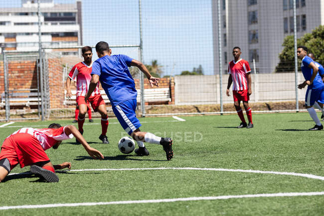 Due squadre multietniche di cinque uomini a squadra giocatori di calcio che indossano una striscia di squadra che giocano una partita in un campo sportivo al sole, affrontando e calciando palla. — Foto stock