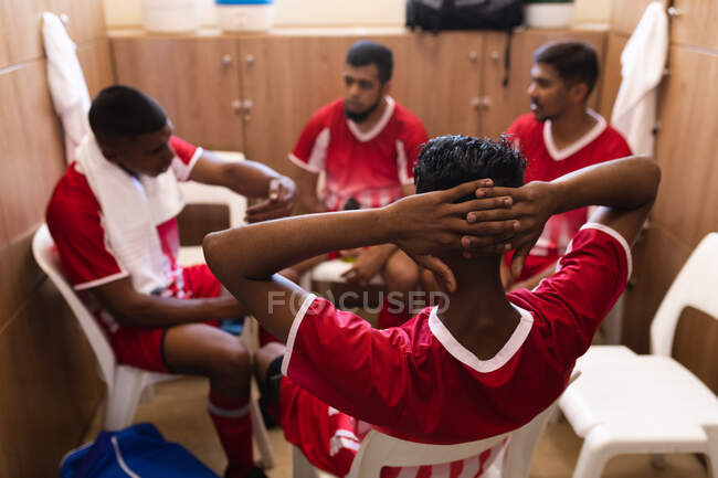Grupo multiétnico de jugadores de fútbol masculino que usan una tira de equipo sentado en el vestuario durante una pausa en el juego, interactuando y hablando. - foto de stock
