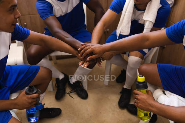 Multi ethnic group of male football players wearing a team strip sitting in changing room during a break in game, hand stacking and holding water bottles. — Stock Photo