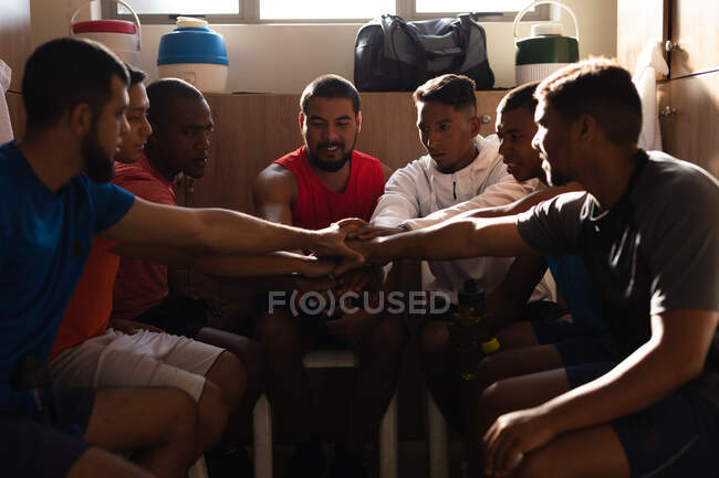 Multi ethnic group of male football players wearing sports clothes sitting in changing room during a break in game, hand stacking and motivating each other. — Stock Photo