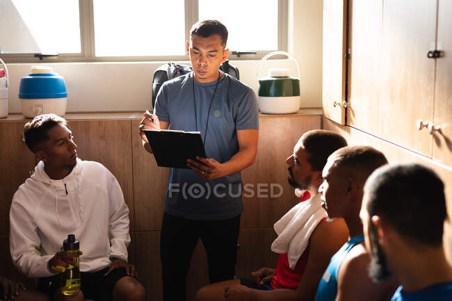 Groupe multi-ethnique de joueurs de football portant des vêtements de sport assis dans un vestiaire pendant une pause dans le jeu, écoutant leur entraîneur instruire tenant bouteille d'eau. — Photo de stock