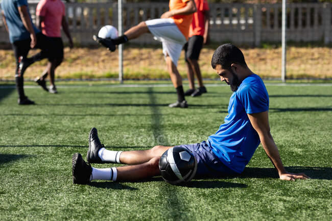 Mixed race male five a side football player wearing sports clothes training at a sports field in the sun, resting sitting with a ball his teammates kicking ball in the background. — Stock Photo