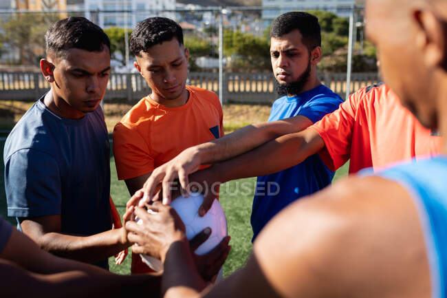 Multi grupo étnico de cinco hombres un lado jugadores de fútbol con entrenamiento de ropa deportiva en un campo de deportes en el sol, de pie mano apilamiento en una pelota motivante antes de un juego. - foto de stock