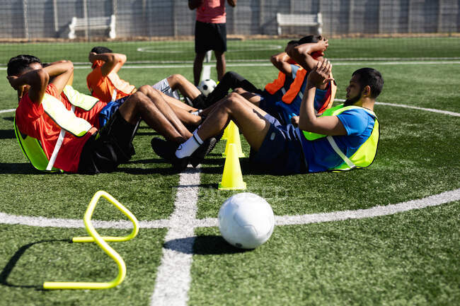 Multi grupo étnico de cinco homens jogadores de futebol um lado vestindo roupas esportivas e coletes de treinamento em um campo de esportes ao sol, aquecendo-se fazendo sentar-se com bola e cones ao lado deles . — Fotografia de Stock