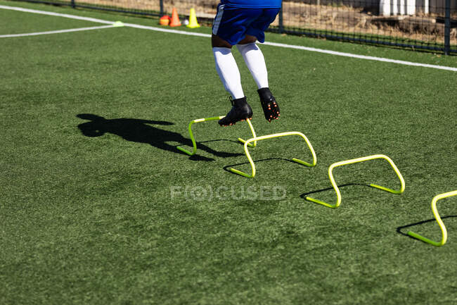 Low section of male football player wearing a team strip training at a sports field in the sun, warming up jumping over hurdles. — Stock Photo