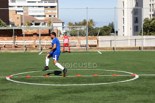 Mixed race male football player wearing a team strip training at a sports field in the sun, warming up jumping over cones another player standing in goal in the background. — Stock Photo