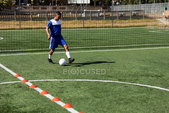 Mixed race male five a side football player wearing a team strip training at a sports field in the sun, warming up tackling with ball. — Stock Photo