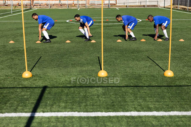 Multi ethnic team of male five a side football players wearing a team strip training at a sports field in the sun, warming up touching ground between cones. — Stock Photo