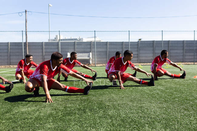 Мультинациональная команда из пяти футболистов, одетых в командную раздевалку на спортивной площадке под солнцем, разогревается, растягиваясь. — стоковое фото