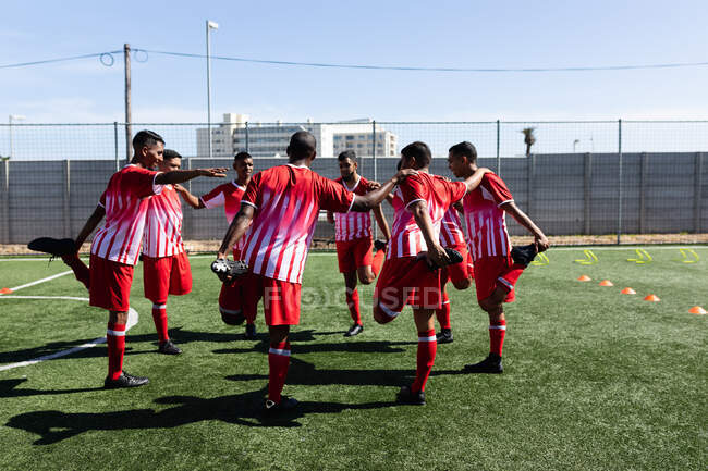 Multi-ethnische Mannschaft von fünf männlichen Fußballern an einer Seite, die auf einem Sportplatz in der Sonne trainieren und sich im Kreis aufwärmen, sich gegenseitig stützen und strecken. — Stockfoto