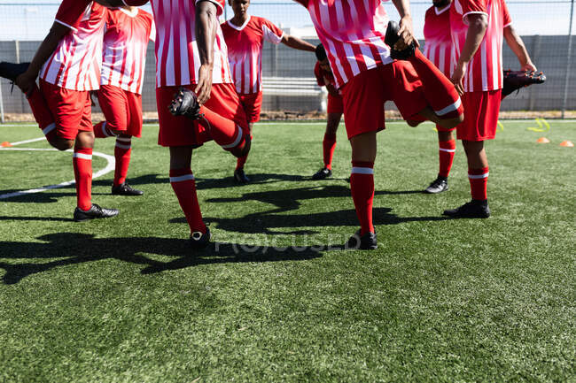 Многонациональная команда из пяти футболистов, одетых в командную раздевалку на спортивной площадке под солнцем, разогревается стоя в кругу, растягиваясь. — стоковое фото