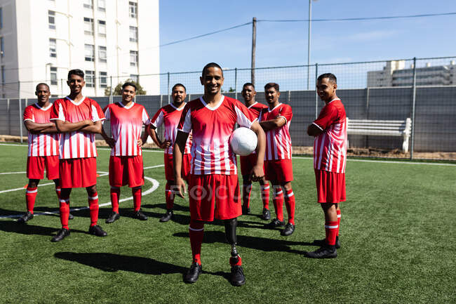 Портрет многонациональной команды мужчин пятерых футболистов, одетых в командную раздевалку на спортивной площадке под солнцем, стоящих, держа мяч, улыбающийся в камеру — стоковое фото