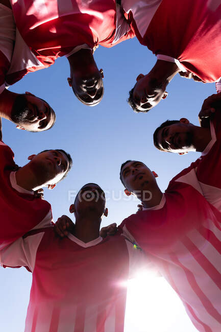 Low angle view of multi ethnic team of male five a side football players wearing a team strip training at a sports field in the sun, standing in huddle motivating each other. — Stock Photo