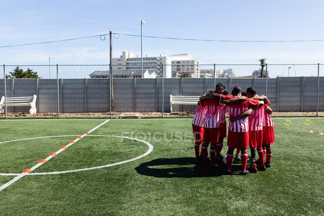 Мультинациональная команда из пяти футболистов, одетых в командную раздевалку на спортивной площадке под солнцем, стоящих в куче мотивирующих друг друга. — стоковое фото