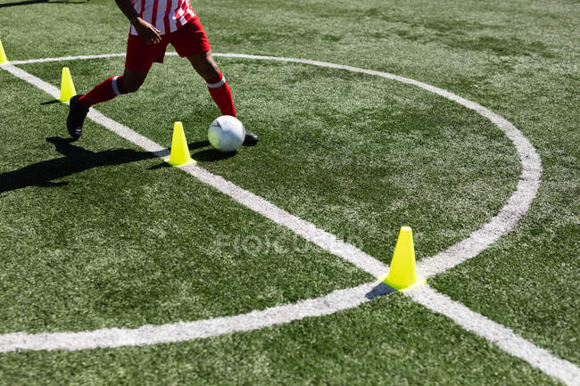 Low section of football player wearing a team strip training at a sports field in the sun, warming up tackling with ball between cones. — Stock Photo