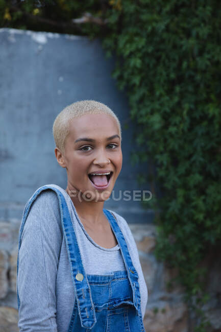 Portrait of mixed race alternative woman with short blonde hair and denim dungarees out and about in the city on a sunny day, smiling to camera. Urban independent woman on the go. — Stock Photo