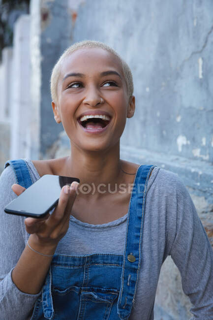 Смешанная расовая альтернативная женщина с короткими светлыми волосами в городе в солнечный день, используя смартфон и смеясь. Городской цифровой кочевник на ходу. — стоковое фото