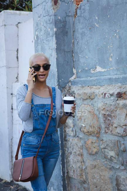 Mujer alternativa de raza mixta con pelo corto y rubio en la ciudad en un día soleado, usando gafas de sol, hablando en smartphone y sosteniendo café para llevar. Nómada digital urbano en movimiento. - foto de stock