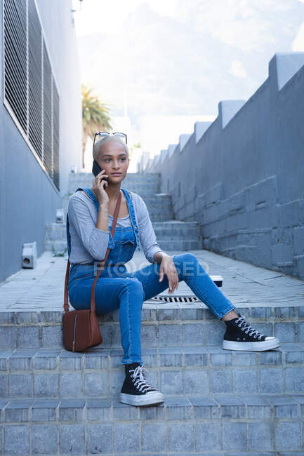 Femme alternative de race mixte avec les cheveux blonds courts dehors et autour dans la ville par une journée ensoleillée, portant des lunettes de soleil et des dungarees de denim, assis sur des étapes à l'aide d'un smartphone. Nomade numérique urbain en déplacement. — Photo de stock