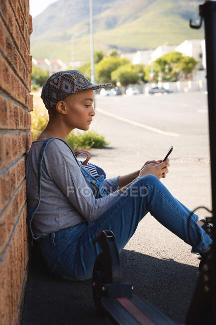 Donna alternativa di razza mista con i capelli corti in giro per la città in una giornata di sole, indossando salopette in denim e un berretto, seduta contro il muro con lo smartphone. Nomade digitale urbano in movimento. — Foto stock
