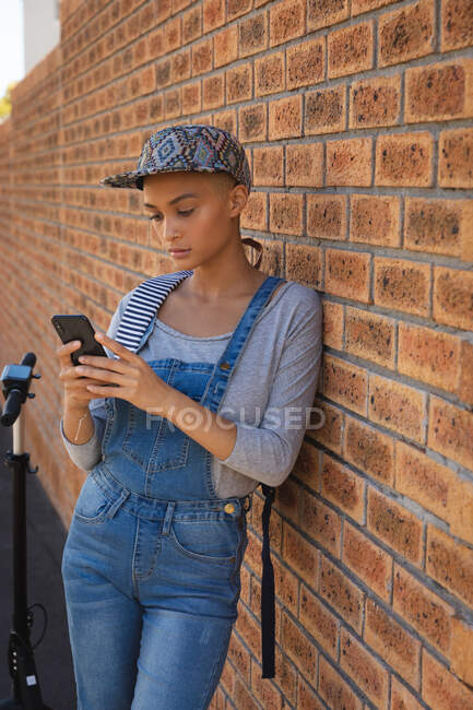 Смешанная расовая альтернативная женщина с короткими светлыми волосами в городе в солнечный день, в джинсовой куртке и кепке, прислонившись к стене с помощью смартфона. Городской цифровой кочевник на ходу. — стоковое фото