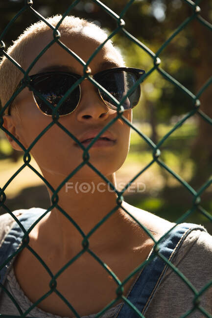 Портрет змішаної раси альтернативної жінки з коротким світлим волоссям на вулиці і близько в місті в сонячний день, одягнені в сонцезахисні окуляри і дивлячись через ланцюгову огорожу. Міська незалежна жінка на ходу . — стокове фото