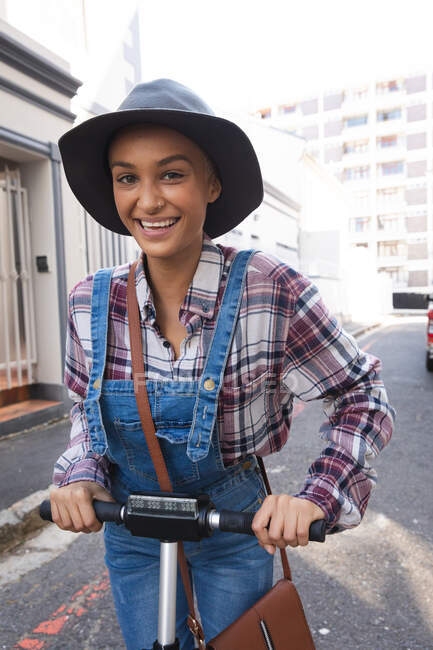 Femme alternative mixte avec les cheveux courts et environ dans la ville par une journée ensoleillée, portant un chapeau et des salopettes, en utilisant un scooter et souriant à la caméra. Nomade numérique urbain en déplacement. — Photo de stock
