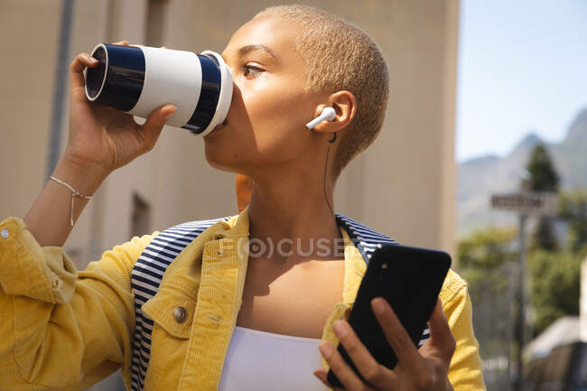 Mujer alternativa de raza mixta con pelo corto y rubio en la ciudad en un día soleado, usando un teléfono inteligente con auriculares inalámbricos y bebiendo un café para llevar. Nómada digital urbano en movimiento. - foto de stock
